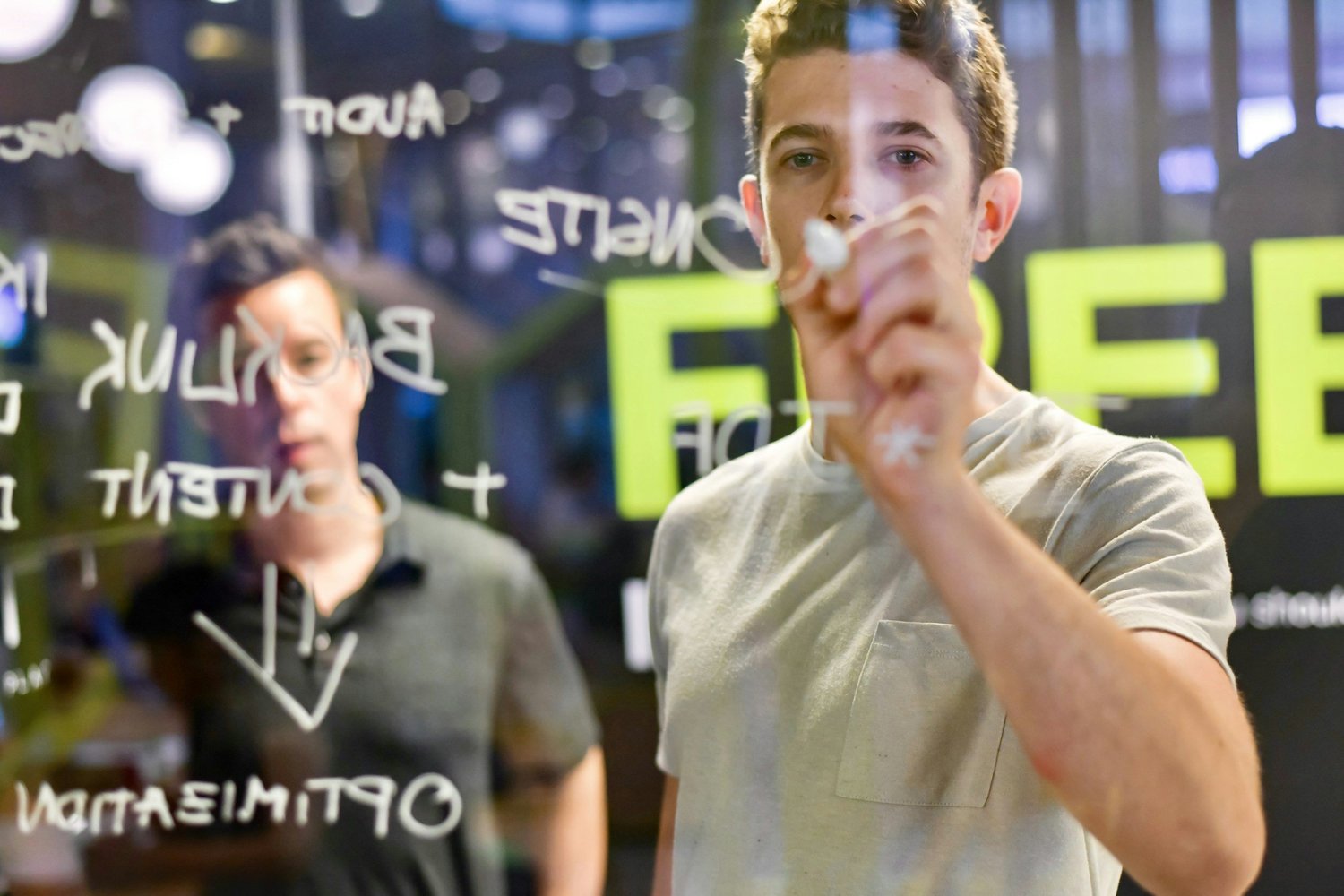 Two young men in a modern office, writing on a glass wall with marker, brainstorming surrounded by brightly lit signs.
