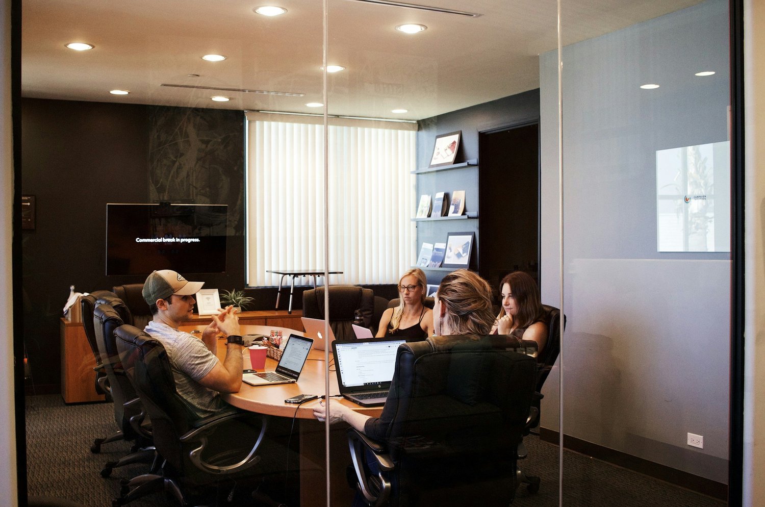 Five people in a conference room working on laptops, with a screen showing 'Commercial break in progress.' on the wall.