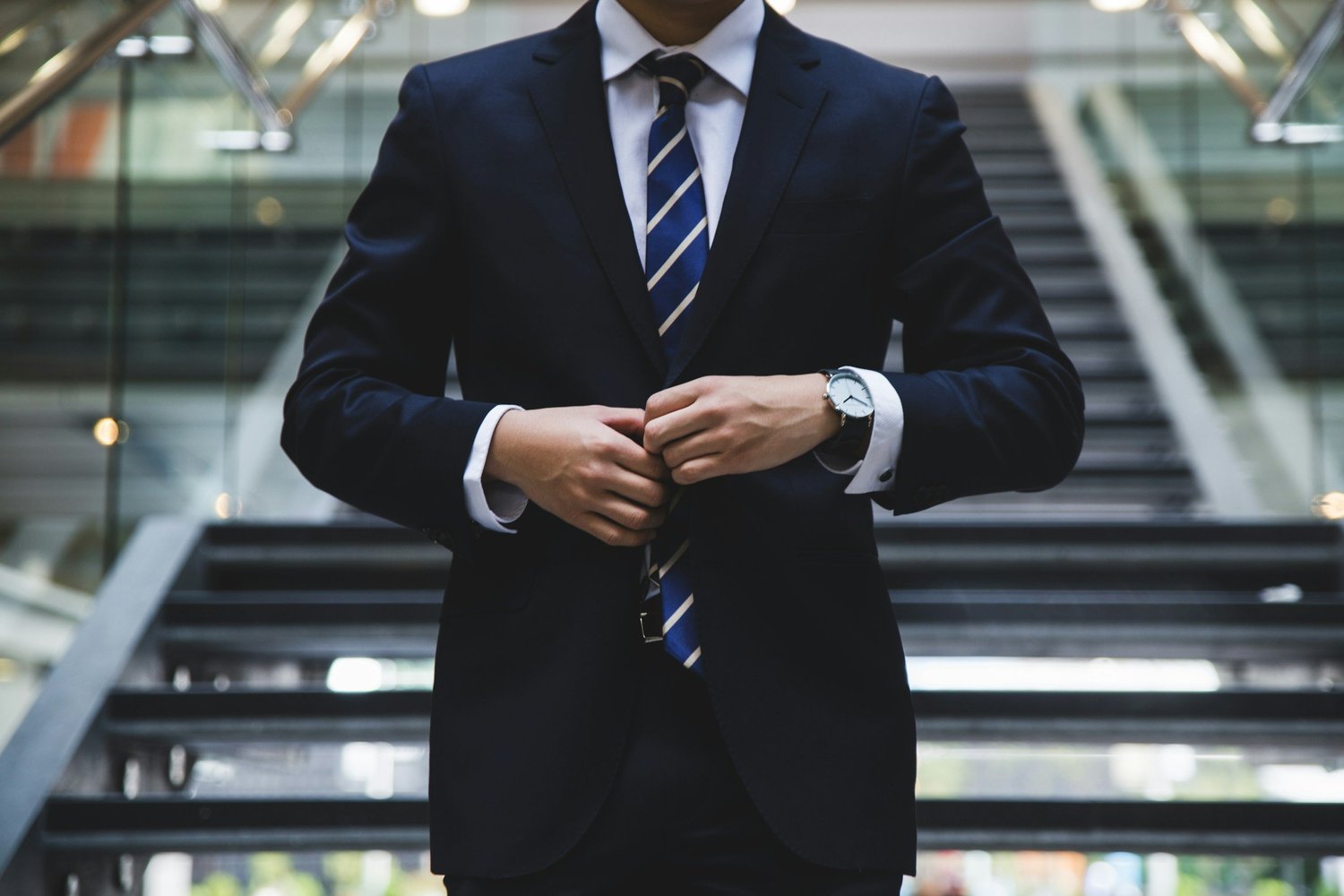 A man wearing a navy blue suit, white shirt, and striped tie adjusting his jacket in front of a staircase in a modern building.