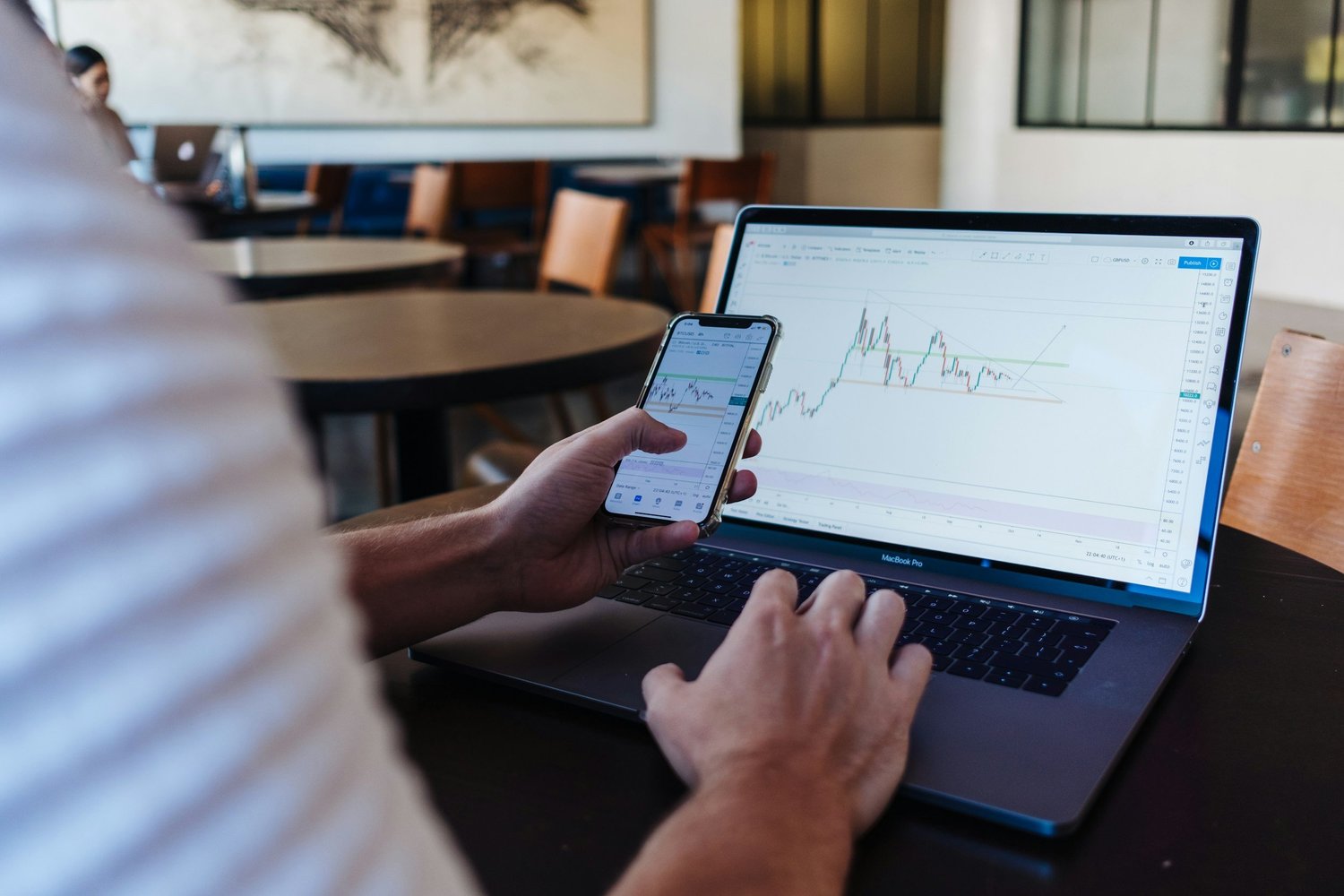 Person using a laptop and smartphone to analyze stock charts in a modern cafe.