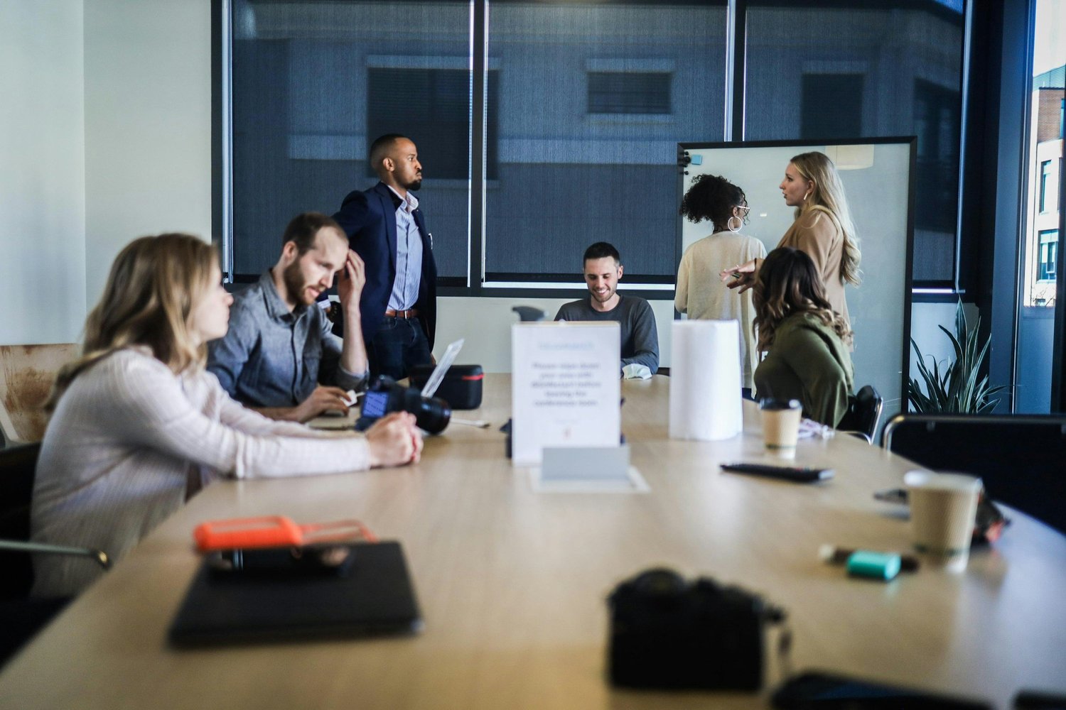 A diverse group of six people in a modern meeting room, some working on laptops and cameras, others talking and engaging in conversation, with large windows and a whiteboard in the background.
