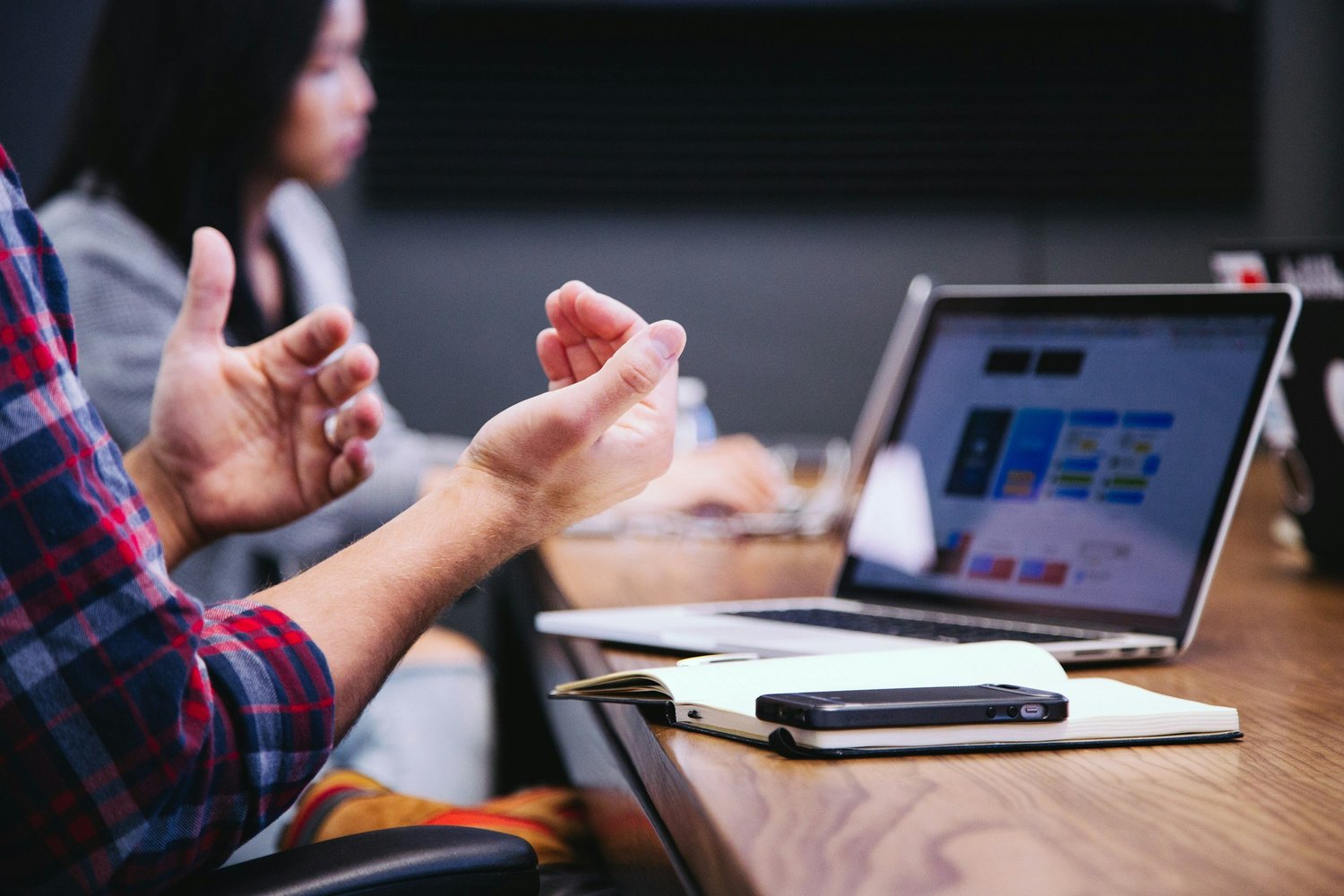 A person in a red and blue checkered shirt gestures with their hands while sitting at a conference table with a laptop, notebook, and smartphone. A woman in a gray blazer is in the background working on a laptop in a meeting room.