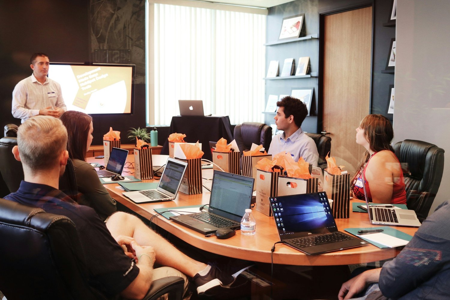 People attending a business meeting or presentation in a conference room, with a presenter at the front and laptops on the table.